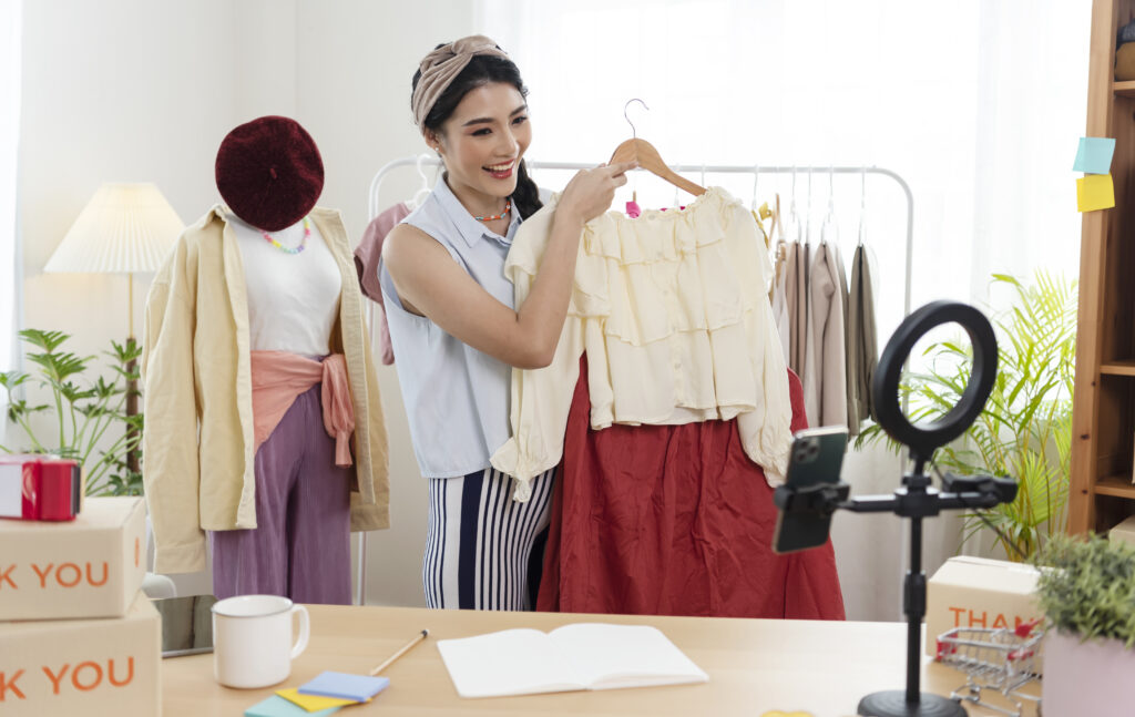 Asian young woman blogger showing clothes in front of smartphone camera while recording vlog video and live streaming at her shop. SME or freelance business online and delivery concept.